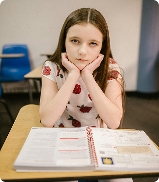 Niña sentada en un escritorio con los codos sobre la mesa y las manos en el rostro, mirando hacia adelante, con libros abiertos frente a ella. Niña sentada en un escritorio con los codos sobre la mesa y las manos en el rostro, mirando hacia adelante, con libros abiertos frente a ella.